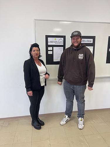 Zwei Personen stehen vor einer Präsentationswand mit Designentwürfen und lächeln in die Kamera.