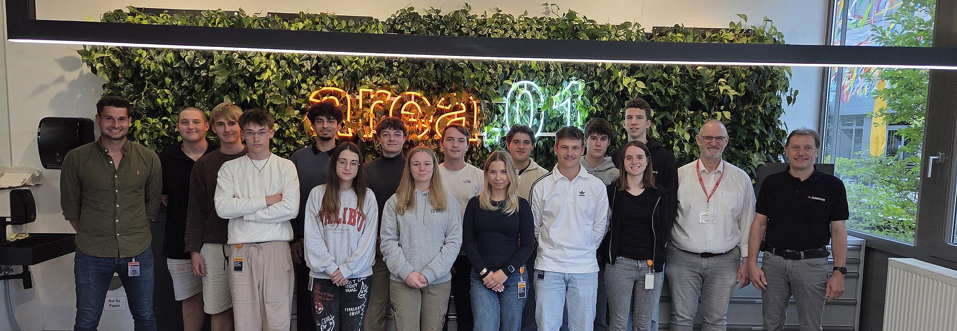 Gruppenfoto einer Ausbildungsgruppe von Aumovio mit Dozenten des Berufsförderungswerks Eckert in einem modernen Schulungsraum. Die Teilnehmenden stehen gemeinsam vor einer begrünten Wand mit leuchtendem Schriftzug „area01“ und blicken in die Kamera.
