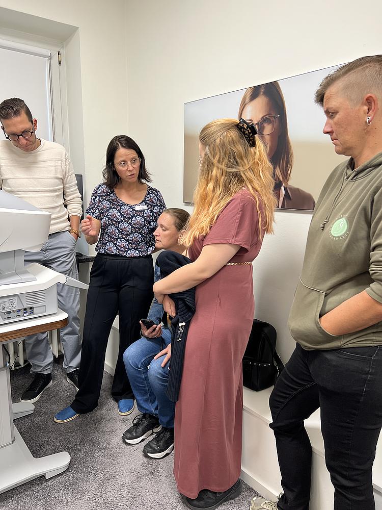 Kleine Gruppe bei einer technischen Gerätevorführung in einem Optikgeschäft mit Erklärungen durch eine Mitarbeiterin.