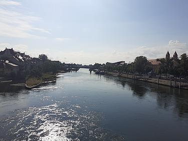 Blick über die Donau in Regensburg bei sonnigem Wetter mit spiegelndem Wasser, Uferpromenade und historischen Gebäuden im Hintergrund.