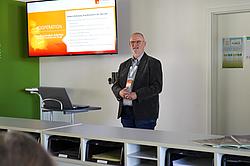 Referent präsentiert Inhalte vor Publikum mit Bildschirm im Hintergrund.