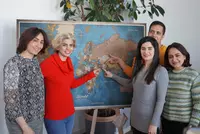 Sechs Personen stehen vor einer großen Weltkarte auf einer Staffelei und lächeln in die Kamera. Einige von ihnen zeigen auf bestimmte Länder auf der Karte, die vermutlich ihre Herkunftsregionen markieren. Im Hintergrund steht eine große grüne Zimmerpflanze.
