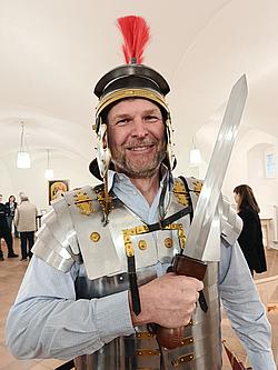 Nahaufnahme eines Mannes mit römischem Helm und Rüstung, der ein Schwert hält.