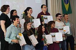 Eine Gruppe von Absolventinnen und Absolventen steht gemeinsam mit ihren Urkunden und Blumen für ein Erinnerungsfoto auf der Bühne.