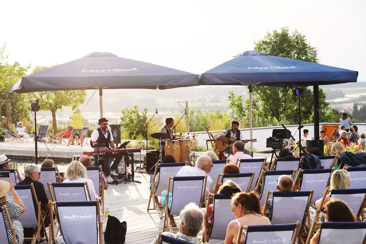 Live-Musik am Eckert Beach bei Sonnenuntergang Drei Musiker treten auf einer Open-Air-Bühne am Eckert Beach auf. Im Vordergrund sitzen Zuschauer in Strandstühlen, während hinter der Bühne eine sommerliche Landschaft im Sonnenlicht zu sehen ist. Zwei große Sonnenschirme mit dem Logo „Eckert Beach“ spenden Schatten über der Bühne.