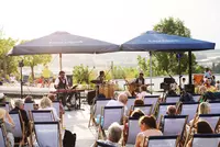 Live-Musik am Eckert Beach bei Sonnenuntergang Drei Musiker treten auf einer Open-Air-Bühne am Eckert Beach auf. Im Vordergrund sitzen Zuschauer in Strandstühlen, während hinter der Bühne eine sommerliche Landschaft im Sonnenlicht zu sehen ist. Zwei große Sonnenschirme mit dem Logo „Eckert Beach“ spenden Schatten über der Bühne.