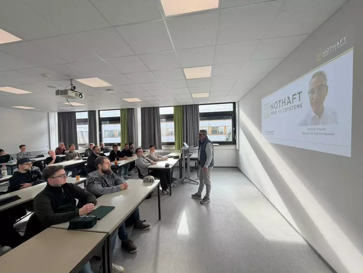 Ein Referent steht vorne in einem modernen Unterrichtsraum und hält einen Fachvortrag vor Vollzeit Studierenden der Weiterbildung zum Heizungs Sanitär und Klimatechniker. Die Teilnehmenden sitzen an Tischen und verfolgen aufmerksam die Präsentation zum Thema Sektorenkopplung und energieeffiziente Gebäudetechnik.