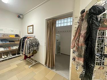 Teilansicht des Fairschenkladens. Man sieht einen Aufssteller mit Schals und CDs, eine Kleiderstange und ein Regal mit weiteren Klamotten. Auf der einen Wandseite befindet sich eine Umkleide. Der Vorhang ist halb offen und man sieht in die leere Kabine hinein.