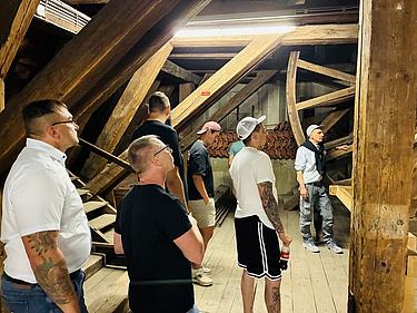 Mehrere Männer stehen in einem historischen Dachstuhl mit massiven Holzbalken und großen Zahnrädern. Ein Führer erklärt die Konstruktion. Die Gruppe betrachtet interessiert die Architektur und Mechanik der alten Holzbauten.