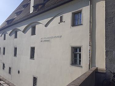 Außenansicht des Gebäudes „Besucherzentrum Welterbe“ in Regensburg mit Fensterfassade und Beschriftung an der Wand
