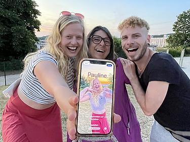 Drei Personen lachen herzlich in die Kamera; im Vordergrund ein Smartphone mit einem scherzhaften Profilbild von „Pauline“, das die Stimmung des Klassentreffens mit Humor unterstreicht. Im Hintergrund: der Eckert Beach bei Sonnenuntergang.