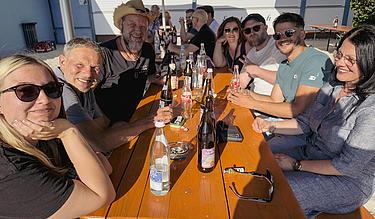 Eine fröhliche Gruppe ehemaliger MG-Teilnehmer:innen sitzt an einem langen Tisch im Freien und lacht in die Kamera. Auf dem Tisch stehen zahlreiche leere und halbvolle Getränkeflaschen. Die Sonne scheint, alle wirken entspannt. Im Hintergrund weitere Personen und das Gebäude des Eckert Beach.