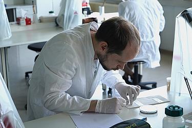 Ein junger Mann im weißen Laborkittel und mit Handschuhen bereitet an einem Labortisch Objektträger für eine mikrobiologische Untersuchung vor.