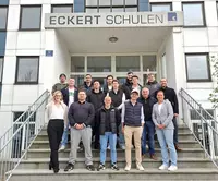 Eine Gruppe von angehenden Industriemeistern steht gemeinsam mit zwei Dozentinnen vor dem Gebäude der Eckert Schulen in Regensburg auf einer Treppe und blickt in die Kamera.