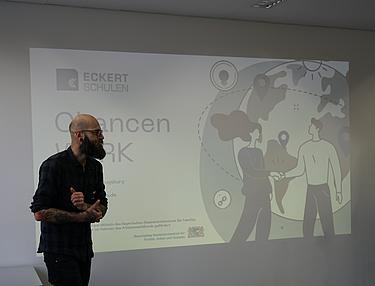 Referent steht vor einer projizierten Präsentation des Projekts ChancenWerk an den Eckert Schulen und spricht zu Teilnehmenden.