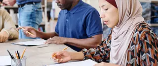 Eine Frau mit Kopftuch und ein Mann sitzen an einem Tisch und bearbeiten konzentriert Prüfungsaufgaben.