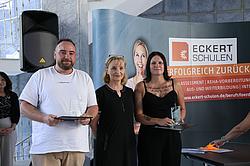 Zwei ausgezeichnete Absolvent:innen stehen mit einer Frau in der Mitte vor einem Roll-Up der Eckert Schulen. Beide halten Urkunden und Glastrophäen in den Händen.