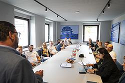 Der Dozent spricht vor der ganzen Gruppe, die an einem langen Tisch sitzt und ihm zuhört. Die Gruppe wirkt sehr interessiert.