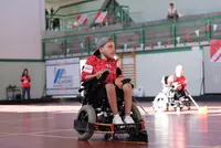 Powerchair-Hockey im Fokus Ein Powerchair-Hockey-Spieler im Rollstuhl mit rotem Trikot auf dem Spielfeld, im Hintergrund weitere Spieler und Zuschauende.