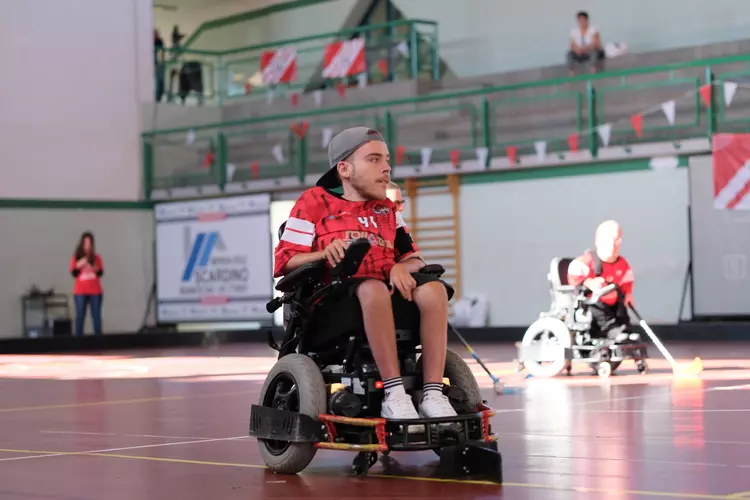 Powerchair-Hockey im Fokus Ein Powerchair-Hockey-Spieler im Rollstuhl mit rotem Trikot auf dem Spielfeld, im Hintergrund weitere Spieler und Zuschauende.