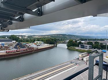 Hafenansicht mit Kränen, Eisenbahnschienen und Lagerflächen am Flussufer.