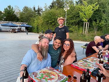 Fünf ehemalige Klassenkameraden sitzen eng beisammen an einem Tisch am Eckert Beach, lachen in die Kamera, vor ihnen Pizza und Getränke; im Hintergrund weitere Teilnehmende in entspannter Atmosphäre.