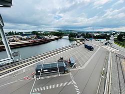 Hafengelände mit Gleisen, Parkplätzen, Containern und einem blauen Lkw, dahinter Fluss, Schrottlager und hügelige Landschaft unter bewölktem Himmel.
