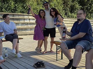 Mehrere Personen stehen und sitzen lachend im Halbkreis auf dem Holzdeck des Eckert Beach. Drei Personen in der Mitte haben sich umarmt und heben fröhlich ihre Getränke in die Luft. Eine weitere Person sitzt vorne rechts und präsentiert augenzwinkernd eine Cola-Mix-Flasche.