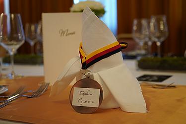 Stoffserviette in Dreiecksform, dekoriert mit einer Medaille am schwarz-rot-goldenen Band, auf der handschriftlich „Giulia Gwinn“ steht; dahinter ein Menüheft auf einem festlich gedeckten Tisch.