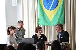 Vier Personen stehen auf einer Bühne vor einer brasilianischen Flagge und überreichen eine Urkunde. Im Vordergrund sitzt ein Publikum und verfolgt die Veranstaltung.