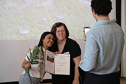 Zwei Frauen posieren für ein Foto, eine hält eine Urkunde und eine Blume, während eine dritte Person das Bild mit einem Smartphone aufnimmt.