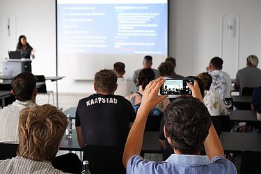 Blick in den Veranstaltungsraum während des Infoabends zum China-Austauschprogramm. Eine Person fotografiert die Präsentation mit dem Smartphone, während Frau Hua Lei vorne spricht.