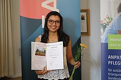 Eine Frau steht vor einem Banner, hält ihre Urkunde und eine Blume und lächelt in die Kamera.