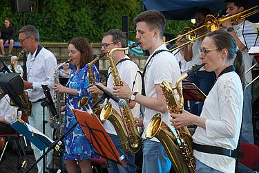 Mehrere Musikerinnen und Musiker spielen Saxophon und Posaune auf einer Open-Air-Bühne. Sie stehen nebeneinander, konzentriert auf ihre Notenständer. Im Hintergrund ist Publikum auf einer Naturtribüne zu sehen.