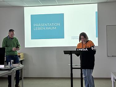 Eine Teilnehmerin präsentiert am Rednerpult ihr Designkonzept, während im Hintergrund die Folie „Präsentation Leben.Raum“ projiziert wird. Neben ihr steht ein Dozent.