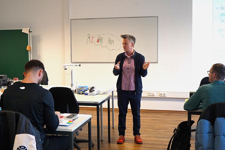 Trainerin Sabine Plettenberg steht vor der Klasse und erklärt gestikulierend einen Lehrinhalt. Hinter ihr hängt ein Whiteboard mit einer gezeichneten Illustration. Vor ihr sitzen Kursteilnehmende an Tischen und hören aufmerksam zu.