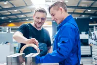 Ein junger Mann in blauer Arbeitskleidung wird von einem Ausbilder in schwarzem Shirt und Latzhose bei der Bearbeitung eines technischen Bauteils angeleitet. Die beiden stehen an einem Werkstück in einer hellen, modernen Werkstattumgebung.