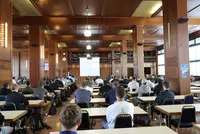 Das Foto zeigt die Bibliothek der Eckert Schulen, in der zahlreiche Teilnehmende der Vollzeit-Weiterbildung „Staatlich geprüfter Techniker“ sitzen. In dem großen, holzvertäfelten Saal stehen Tische und Stühle in Reihen. Die Teilnehmenden blicken nach vorne zu einer Leinwand, auf der eine Präsentation gezeigt wird. Dort begrüßt Geschäftsführer und Schulleiter Markus Johannes Zimmermann die neuen Vollzeit-Techniker offiziell zum Start ihres Lehrgangs.