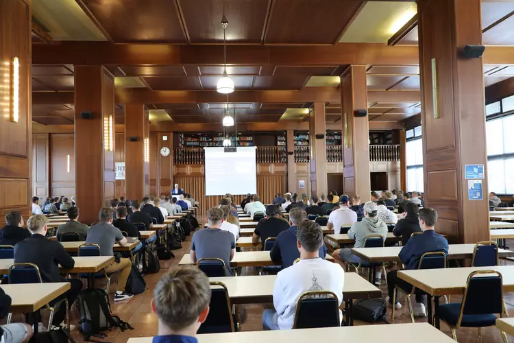 Vollzeit-Technikerstart in der Bibliothek der Eckert Schulen: Begrüßung durch Geschäftsführer und Schulleiter Markus Johannes Zimmermann Das Foto zeigt die Bibliothek der Eckert Schulen, in der zahlreiche Teilnehmende der Vollzeit-Weiterbildung „Staatlich geprüfter Techniker“ sitzen. In dem großen, holzvertäfelten Saal stehen Tische und Stühle in Reihen. Die Teilnehmenden blicken nach vorne zu einer Leinwand, auf der eine Präsentation gezeigt wird. Dort begrüßt Geschäftsführer und Schulleiter Markus Johannes Zimmermann die neuen Vollzeit-Techniker offiziell zum Start ihres Lehrgangs.