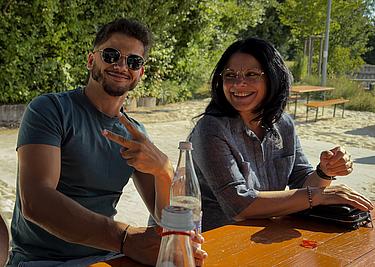 Zwei ehemalige Teilnehmer:innen der MG-Klasse sitzen an einem Tisch am Eckert Beach. Der junge Mann zeigt ein Peace-Zeichen und lächelt in die Kamera, die Frau daneben lacht herzlich. Leere Glasflaschen und persönliche Gegenstände liegen auf dem Tisch, im Hintergrund Bänke, Sandfläche und grüne Bäume.