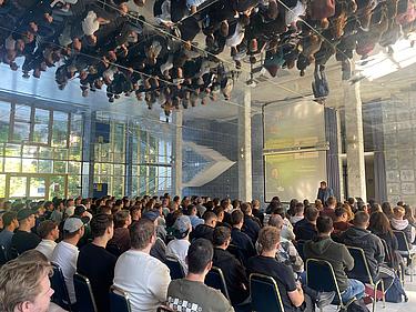 Das Bild zeigt eine große Gruppe von Studierenden, die im Prüfungszentrum der Eckert Schulen Platz genommen hat. Vorne am Podium spricht ein Dozent zur Einführung, während die Präsentation an die Wand projiziert wird. Die Spiegeldecke des Raumes spiegelt die gesamte Szene eindrucksvoll wider.
