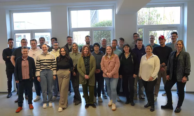 Eine Gruppe von rund 25 Personen steht in einem hellen Unterrichtsraum vor großen Fenstern und blickt lächelnd in die Kamera. Vorne in der Gruppe steht Trainerin Sabine Plettenberg. Die Teilnehmenden wirken motiviert und bunt gemischt.