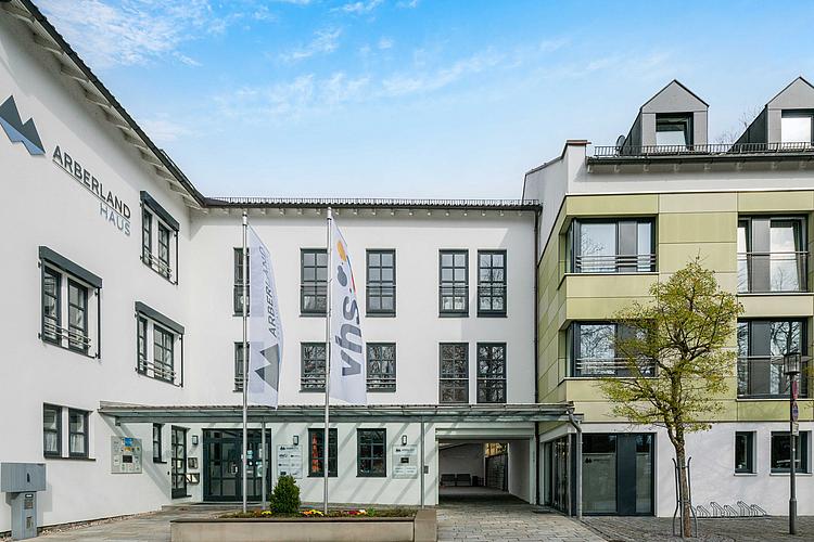 Barrierefreier Eingangsbereich des Arberlandhauses in Regen mit moderner Glasüberdachung, zwei wehenden Fahnenmasten mit den Logos von ARBERLAND und VHS, weißer Gebäudefassade mit schwarzen Fensterrahmen und angebautem grünlichem Flügel auf der rechten Seite. Links am Gebäude ist das ARBERLAND-Logo zu sehen.