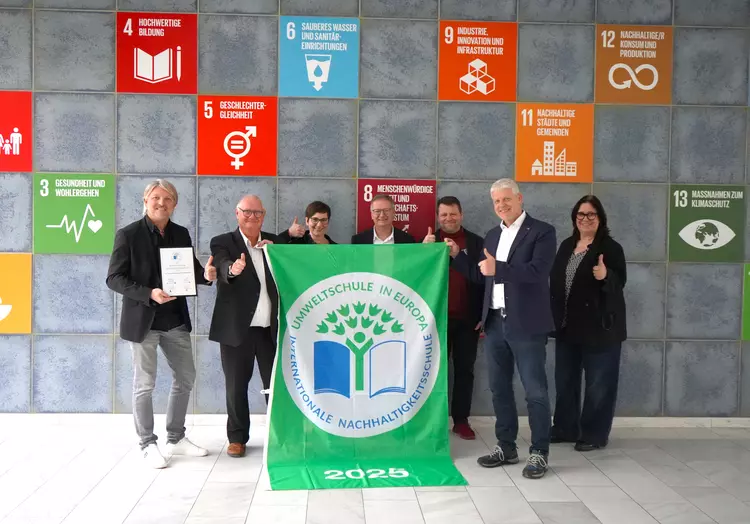 Auszeichnung Umweltschule 2025 Dr. Eckert Akademie Mitarbeitende der Dr. Eckert Akademie stehen vor einer Wand mit Symbolen der Nachhaltigkeitsziele und halten die grüne Flagge „Umweltschule in Europa – Internationale Nachhaltigkeitsschule 2025“.