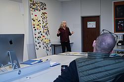 Dozentin erläutert Projektergebnisse vor einer Pinnwand mit kreativen Arbeitsmaterialien und Ideensammlung.