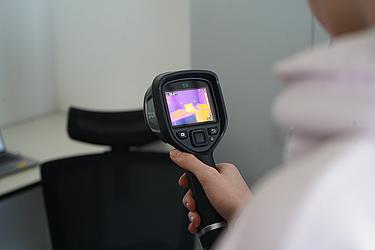 Eine Hand hält eine Wärmebildkamera, deren Display farbige Temperaturunterschiede anzeigt.