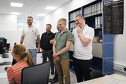 Eine Gruppe von Personen stehen um eine junge Frau, die an einem Computer arbeitet. Alle Beteiligten wirken interessiert und lachen freundlich.