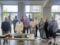 Gruppe von Teilnehmenden steht gemeinsam in einem hellen Raum hinter einem Tisch mit Arbeitsmaterialien; eine Person trägt einen weißen Laborkittel.