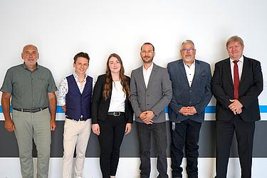 Gruppenbild von 14 Personen, darunter Studierende, Lehrkräfte und Projektbeteiligte. Sie stehen nebeneinander vor einer weißen Wand und blicken in die Kamera.
