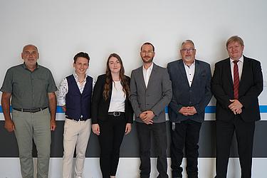 Gruppenbild von 14 Personen, darunter Studierende, Lehrkräfte und Projektbeteiligte. Sie stehen nebeneinander vor einer weißen Wand und blicken in die Kamera.